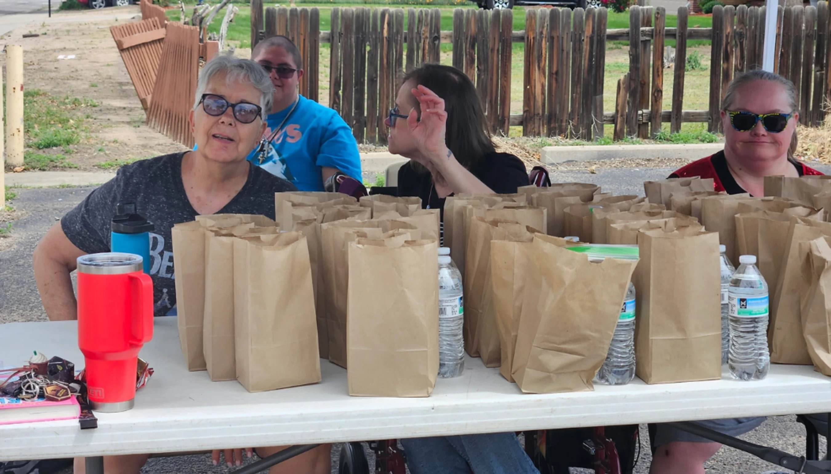 A group of four people sit at a long white table outdoors, which is filled with many brown paper bags and bottles of water, ready for distribution.