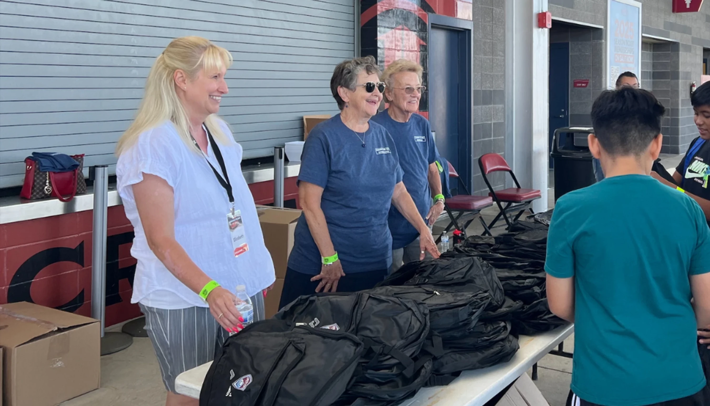 Three smiling women stand behind a table piled with black backpacks, handing them to young boys at an event.