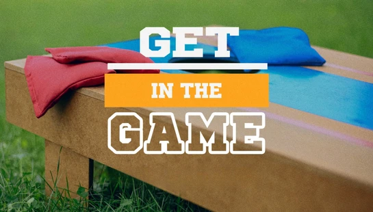 A cornhole board on a green lawn with red and blue bean bags. Text overlay reads, 'Get In The Game' in a white, athletic-style font.