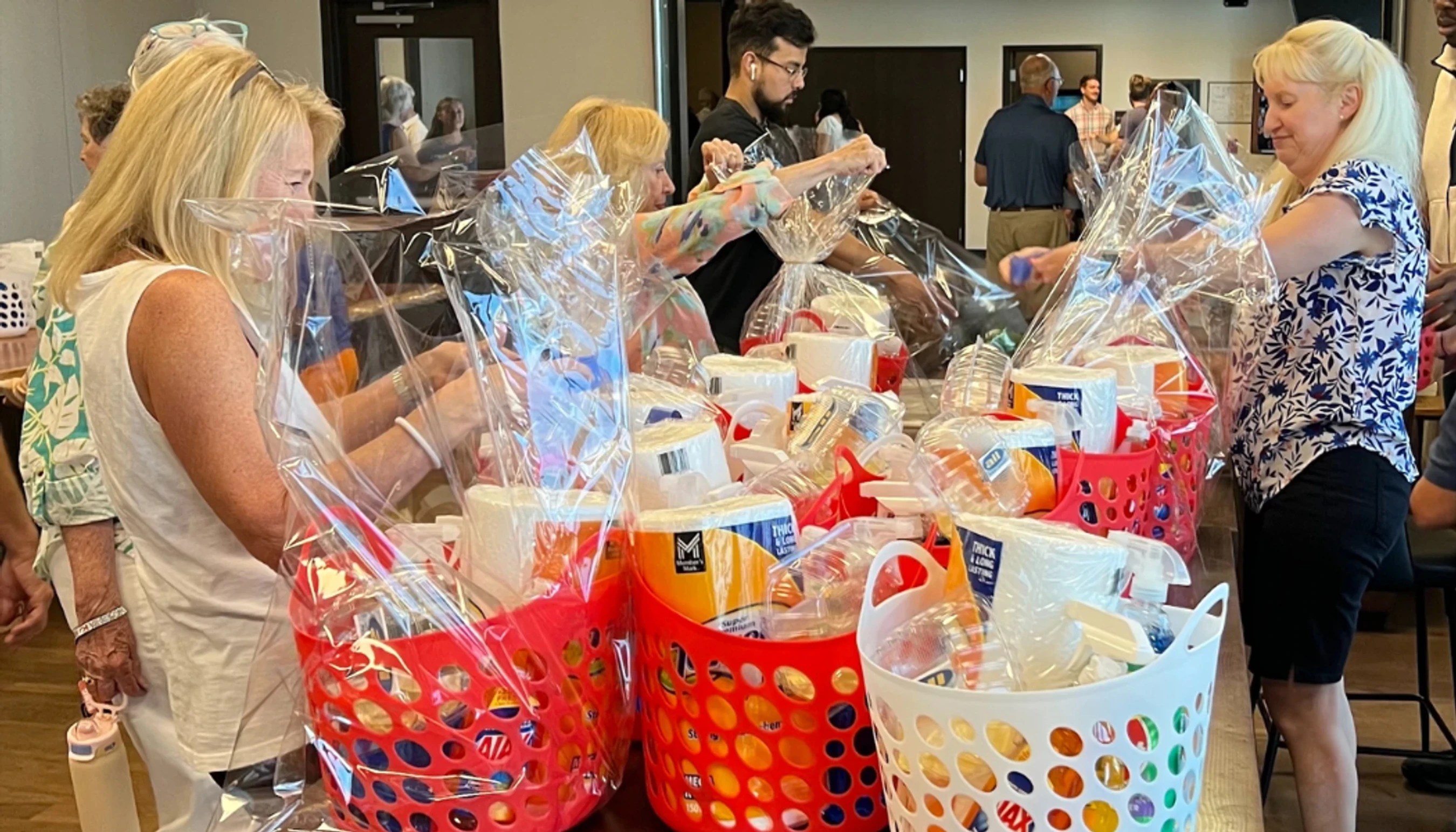 A group of people stand around a table assembling care packages, filling colorful baskets with household supplies and wrapping them in clear plastic.