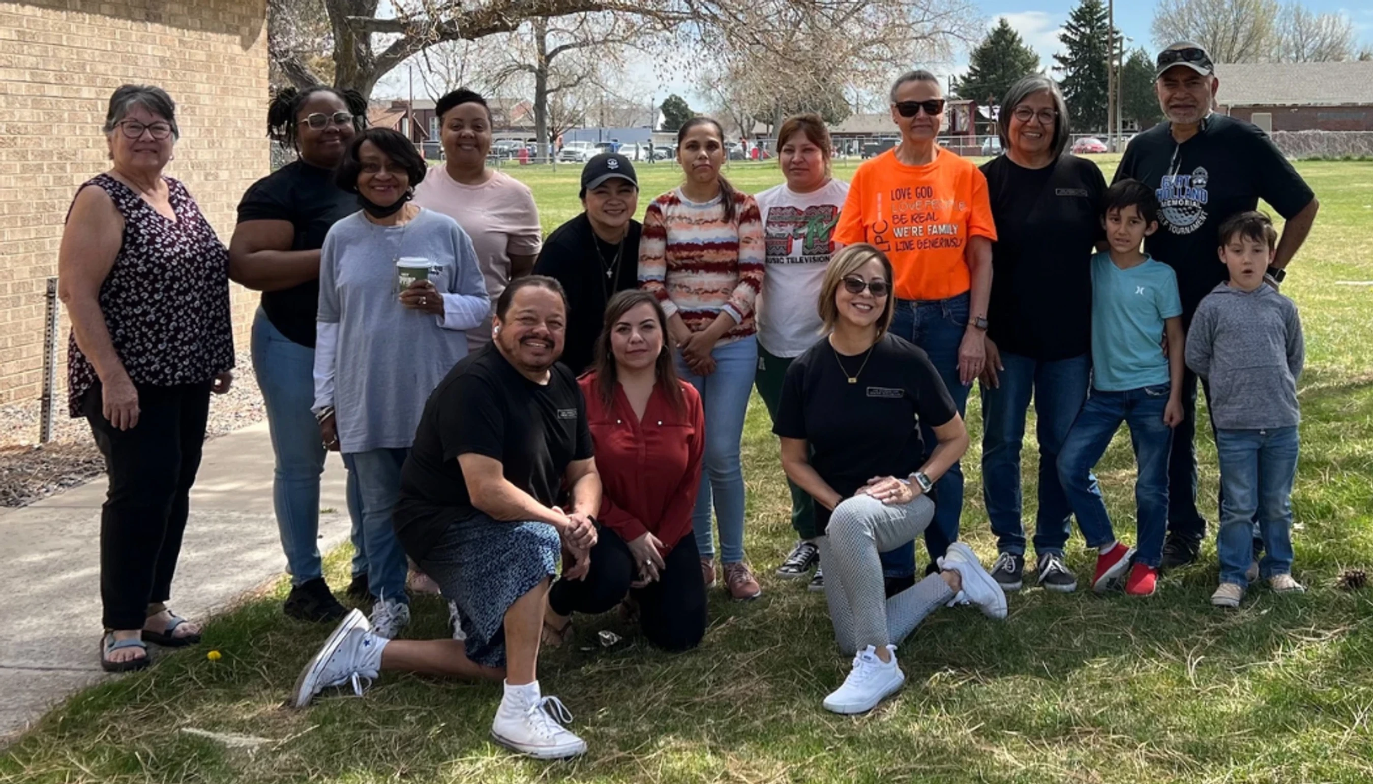 A diverse group of adults and two young boys posing together for a group photo on a grassy lawn in front of a brick building on a sunny day.