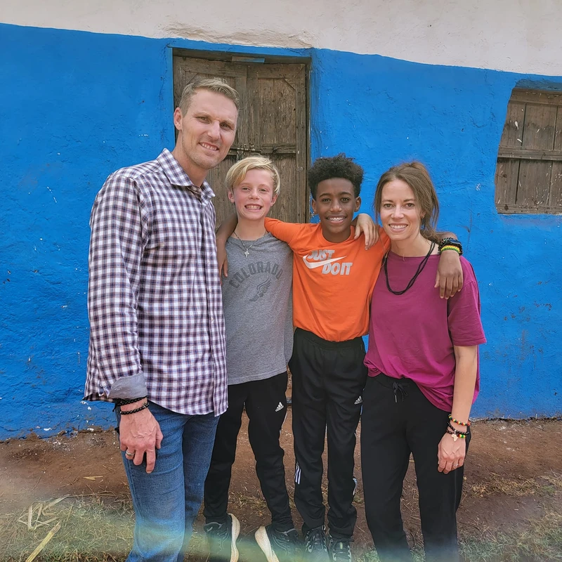 A family of four, two adults and two boys, stand together smiling for a photo in front of a rustic building with a bright blue painted wall.