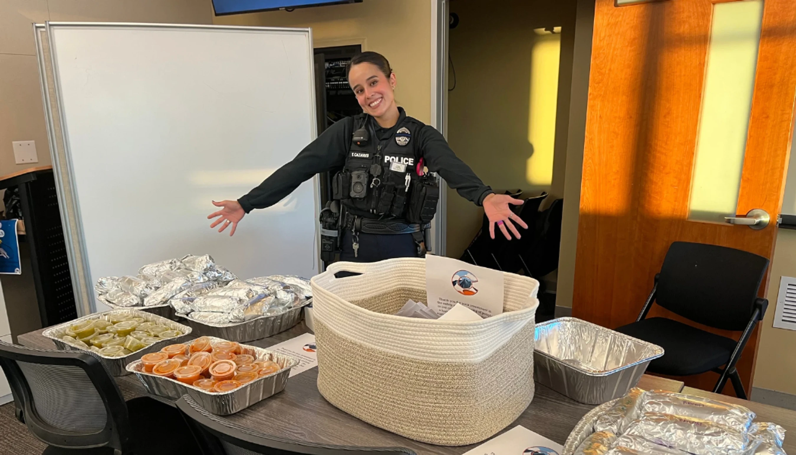 A smiling female police officer in uniform stands with her arms outstretched behind a table laden with burritos and salsas.