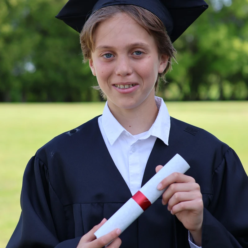 A young person with braces smiles while wearing a black graduation cap and gown. They are holding a diploma tied with a red ribbon outdoors.