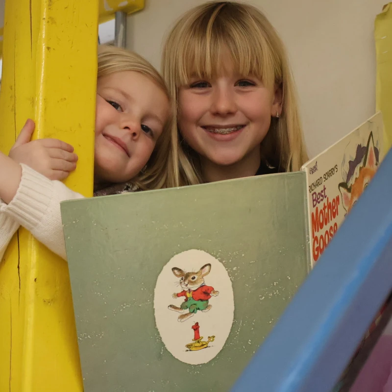 Two young blonde sisters smile while holding a 'Best Mother Goose Ever' book. The older girl has braces, and the younger one peeks from behind a yellow post.
