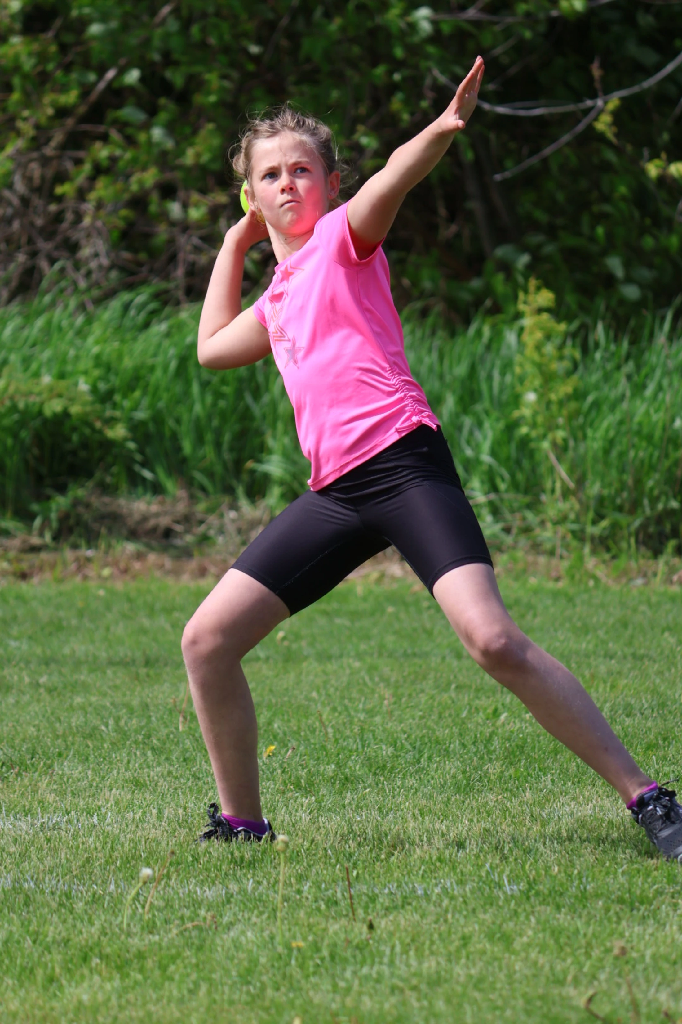 A young girl in a bright pink shirt and black shorts stands on a grassy field, her face focused as she winds up to throw a yellow ball.