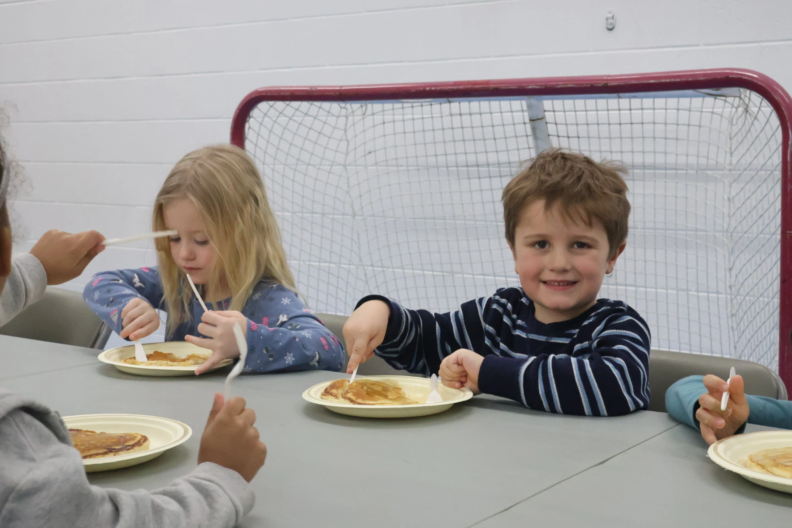 A young boy in a striped shirt smiles at the camera while eating pancakes at a table with other children. A hockey net is visible in the background.