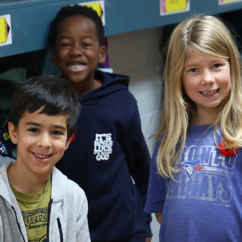 Three diverse elementary school-aged children, two boys and a girl, stand close together in front of classroom cubbies and smile for the camera.