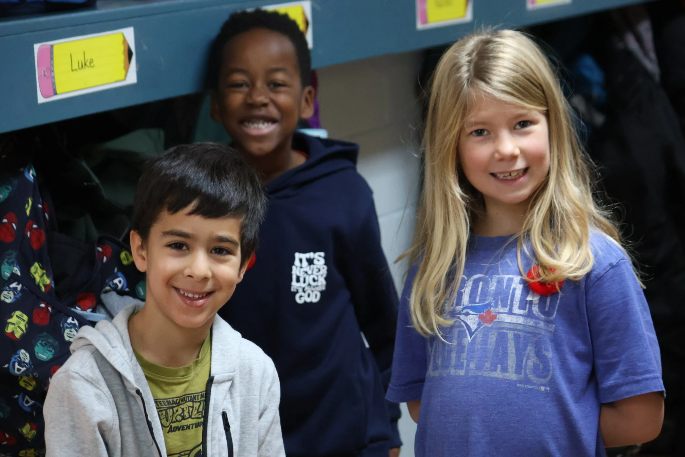 Three diverse elementary school-aged children, two boys and a girl, stand close together in front of classroom cubbies and smile for the camera.