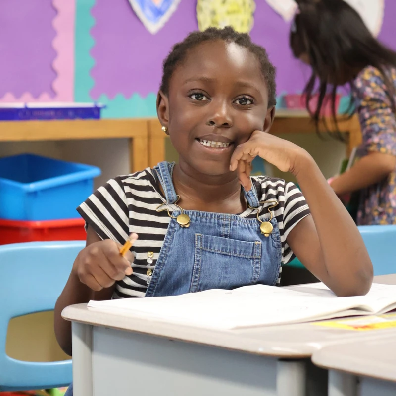 A young Black girl with braids and braces, wearing denim overalls, smiles at the camera while sitting at her desk in a colorful classroom and holding a pencil.