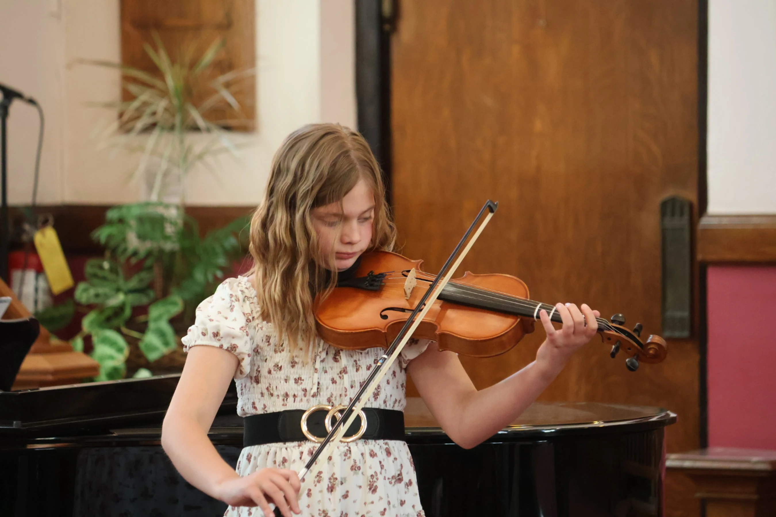 A young girl with wavy blonde hair in a floral dress concentrates as she plays the violin during an indoor performance.
