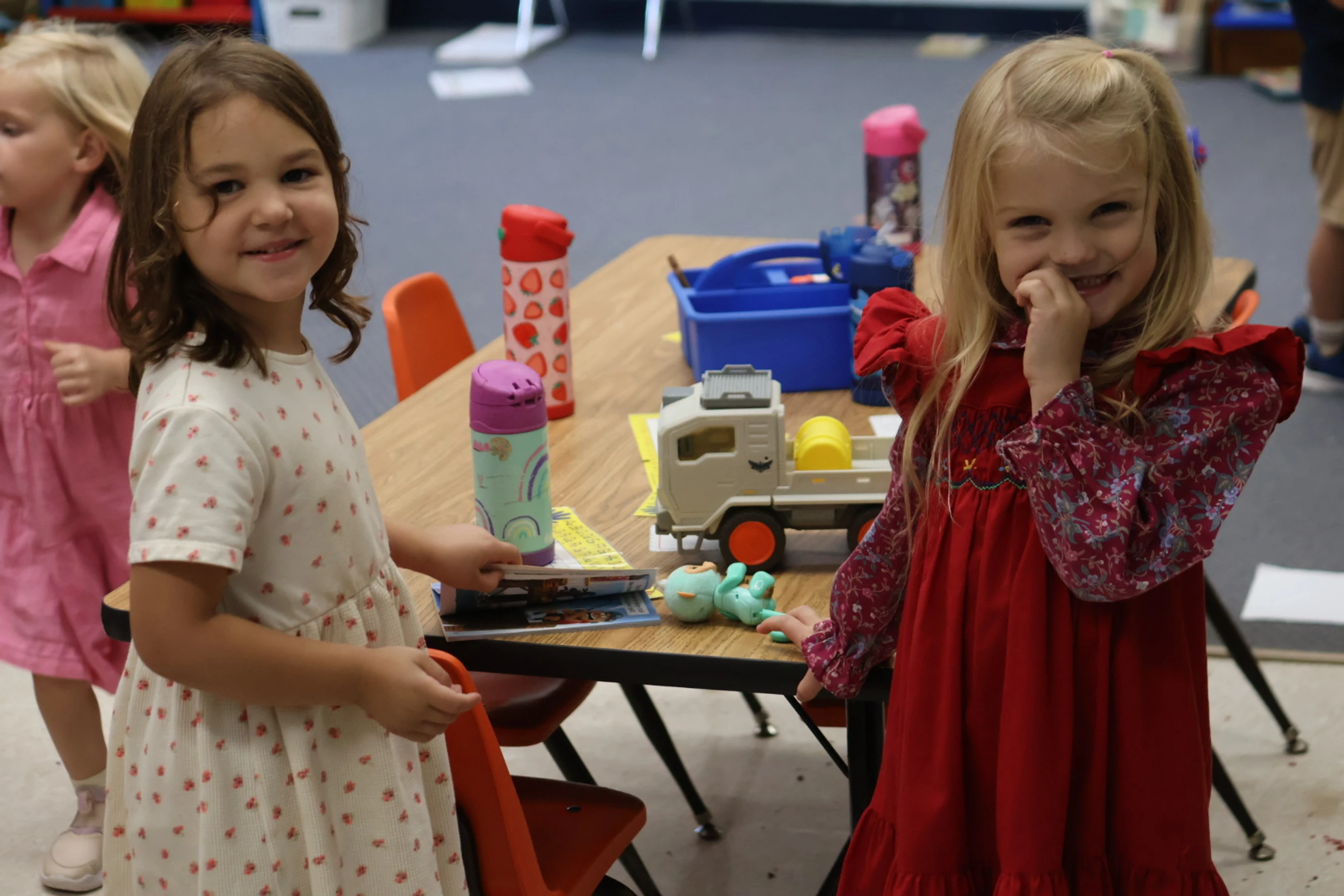 Two young girls, one in a white floral dress and one in a red dress, stand by a table with toys in a classroom and smile at the camera.