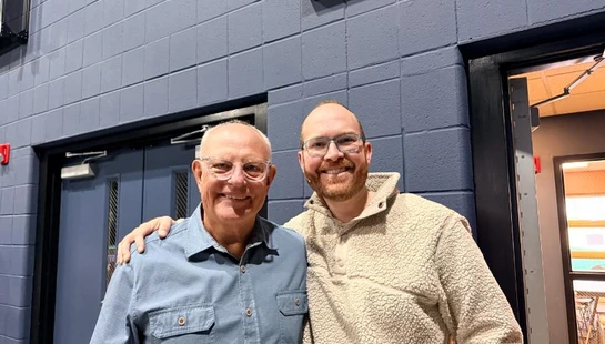 An older man in a blue shirt and a younger man in a fleece pullover smile for the camera, standing arm-in-arm in a hallway with a blue brick wall.