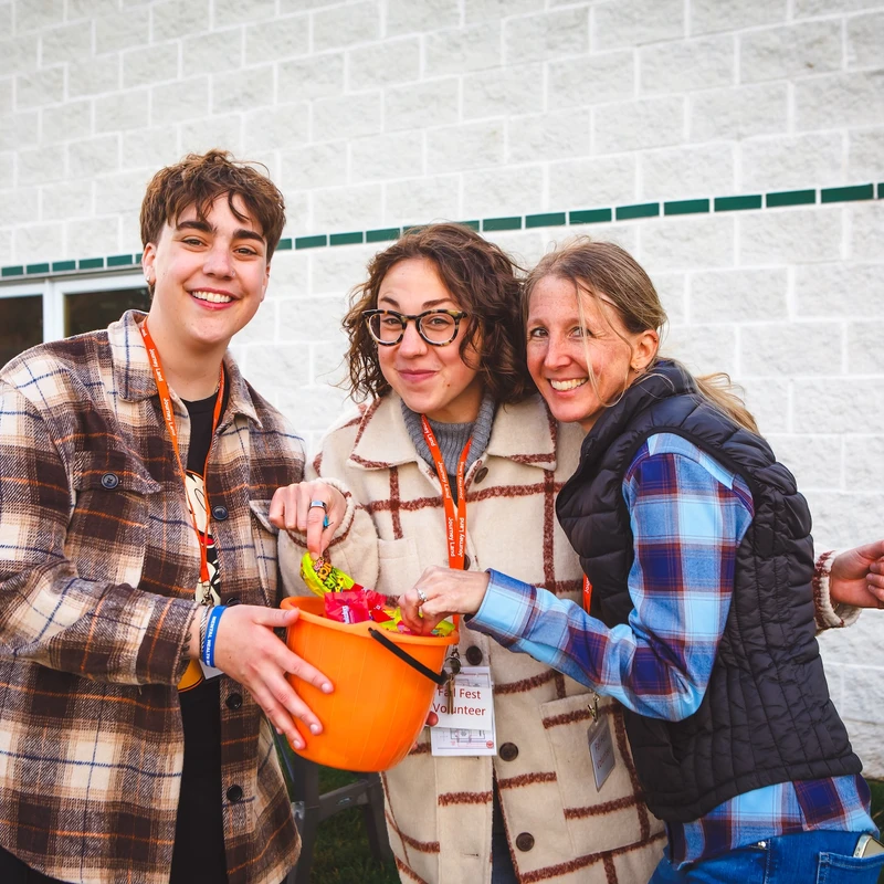 A young person and two women, all smiling volunteers, pose for a photo while holding an orange bucket filled with candy during a fall festival.