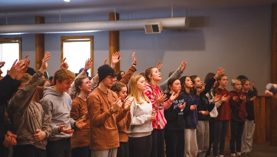 A large group of young people standing indoors, singing with their hands raised in an act of worship during a gathering or service.