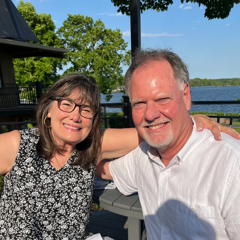 A smiling man and woman pose for a photo on an outdoor patio. The woman has her arm around the man's shoulder. A lake and trees are in the background.