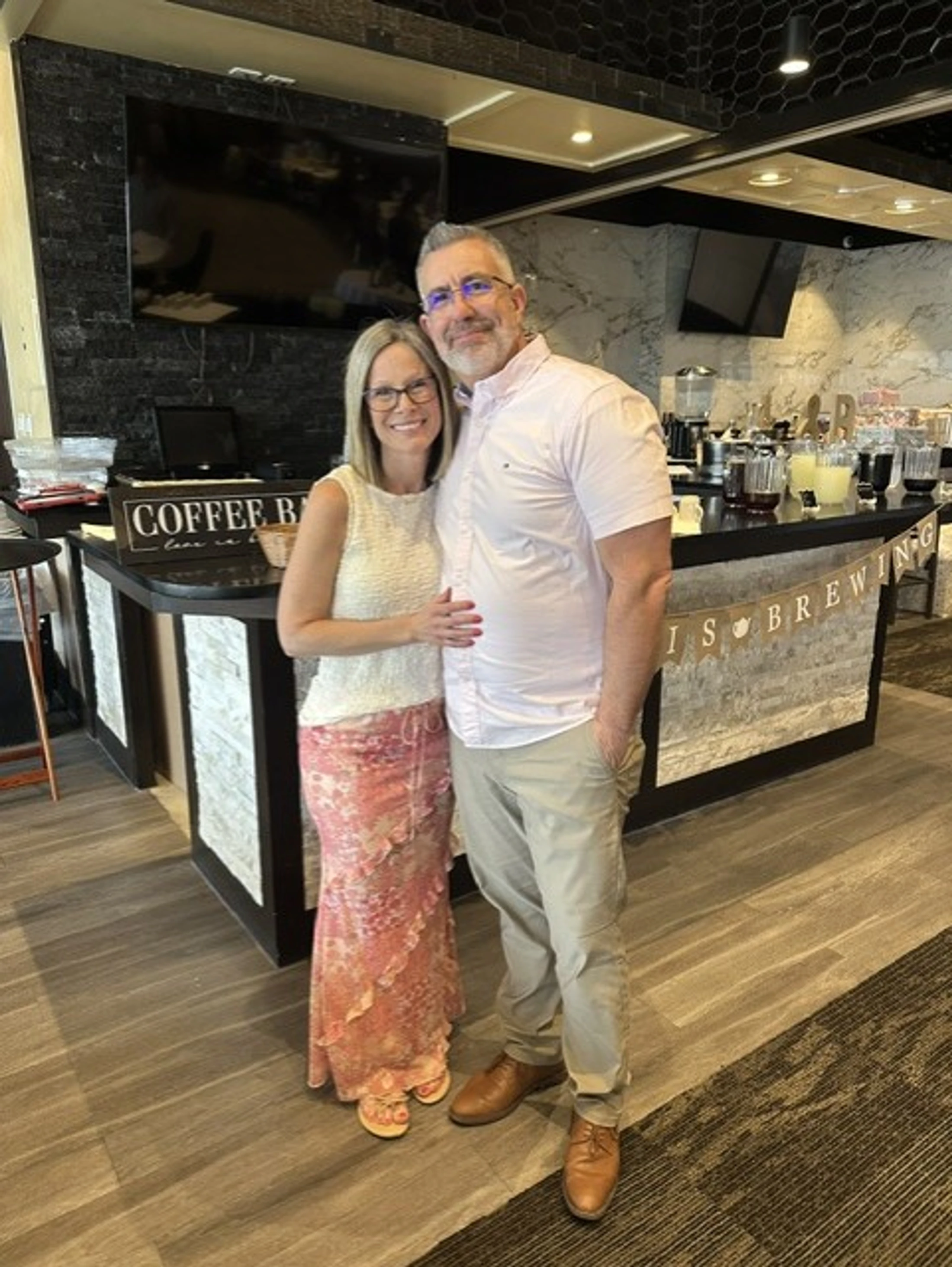 A smiling couple poses for a photo at an indoor event. The man has his arm around the woman as they stand in front of a modern-looking coffee bar.