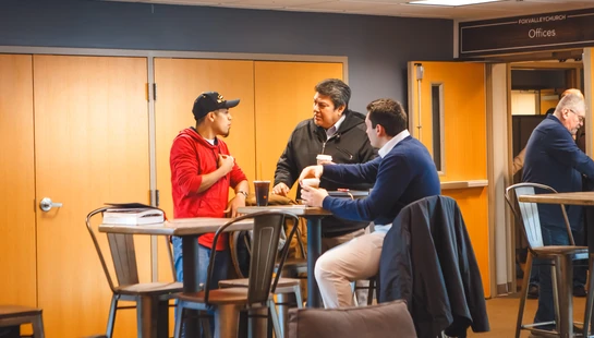 Three men in casual clothes gather around a high-top table, talking and drinking coffee in the lobby of the Fox Valley Church.