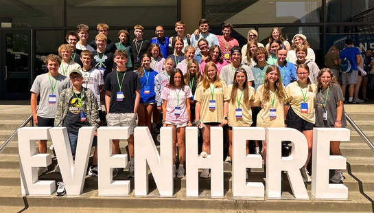 A large group of teenagers wearing lanyards pose for a photo on steps behind large, white, 3D block letters that spell out the word EVENHERE.