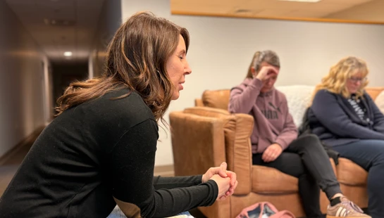 A woman with brown hair sits in profile, speaking seriously with her hands clasped. In the background, two other women listen on a couch, looking somber.
