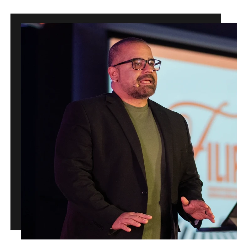 A man with glasses, a beard, and a microphone, wearing a black blazer and green shirt, speaks and gestures while giving a presentation on stage.