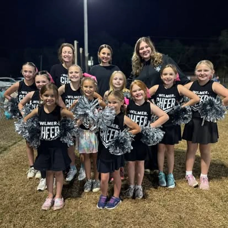 A youth cheerleading squad and their coaches posing for a group photo on a grassy field at night. The girls wear black uniforms and hold silver pom-poms.