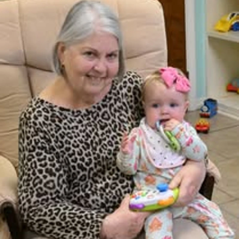 A smiling older woman with grey hair sits in a beige armchair, holding a baby girl in a patterned onesie and a pink bow on her lap.