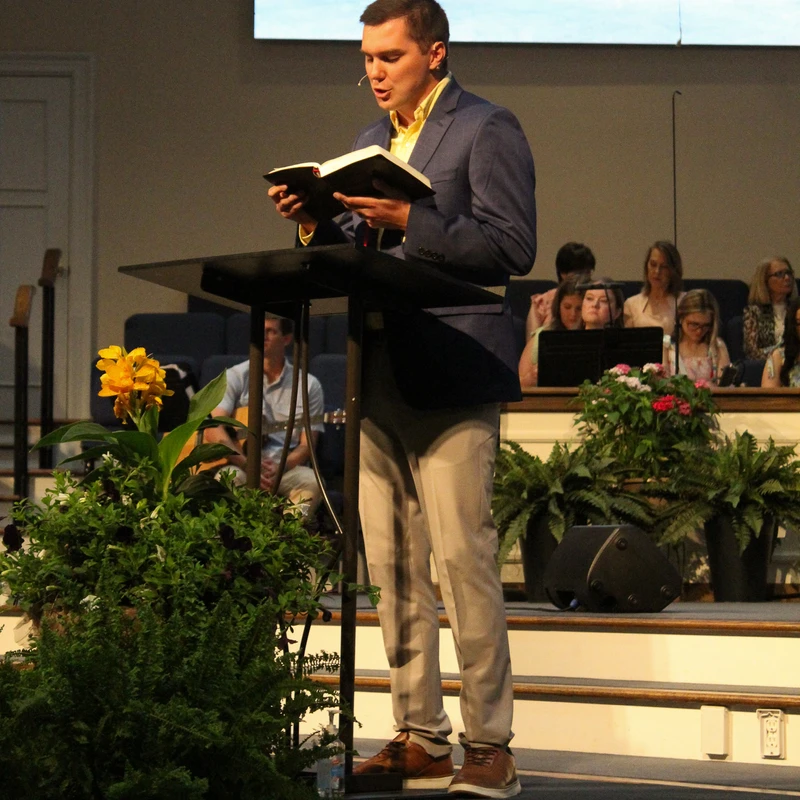 A young man in a blue blazer stands at a lectern on a church stage, reading from a book. A scripture verse is projected on a screen behind him.