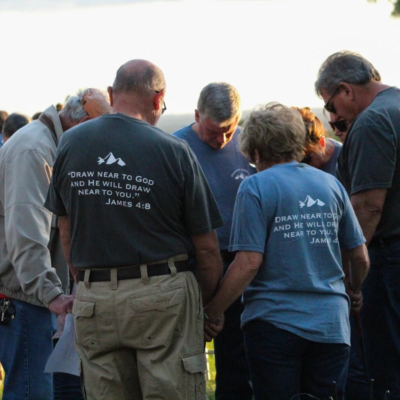 A group of people stand in a circle outdoors, holding hands and bowing their heads in prayer. Many wear shirts with a Bible verse on the back.
