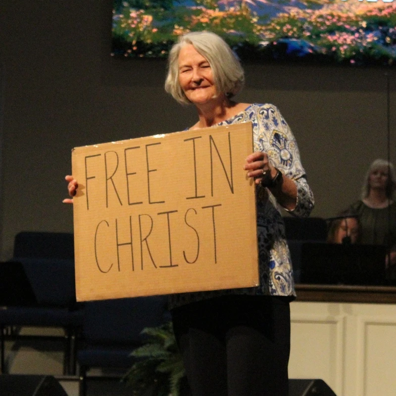 A smiling woman with gray hair stands on a church stage, holding a handmade cardboard sign that reads 'FREE IN CHRIST' in black marker.