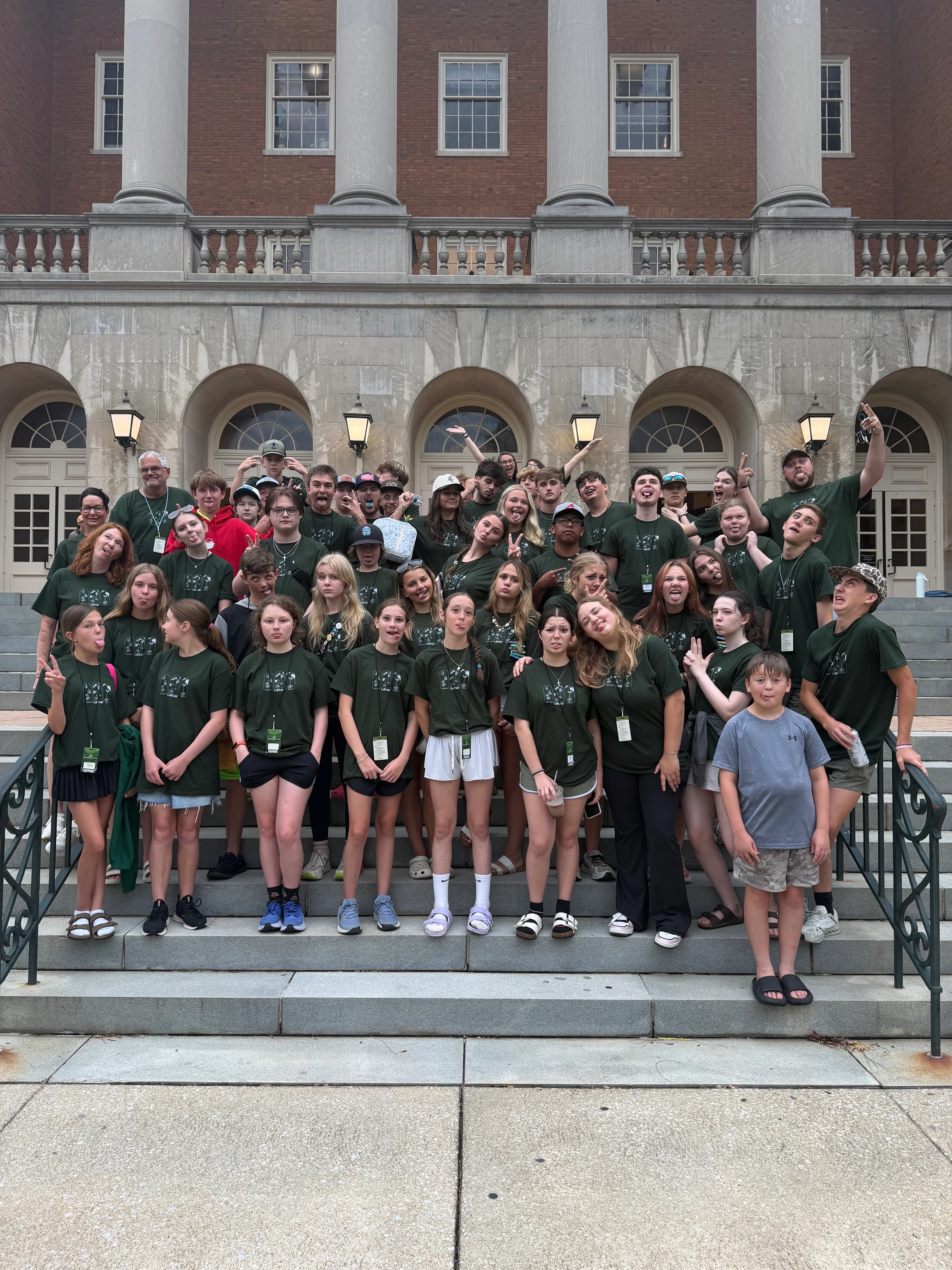 A large group of young people and adults in matching green t-shirts make funny faces while posing for a photo on the steps of a grand, columned building.