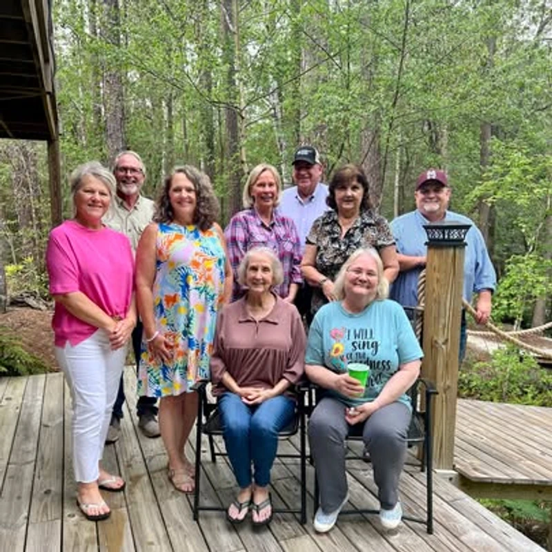 A group of nine smiling adults, men and women, pose together for a photo on a wooden deck with a lush green forest in the background.