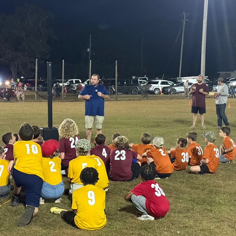 A male coach speaks into a microphone, addressing a large group of children in sports jerseys who are sitting on a grassy field at night.