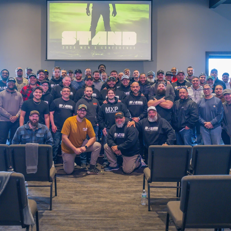A large group of diverse men posing for a photo indoors in front of a projector screen that reads 'STAND 2026 Men's Conference'.