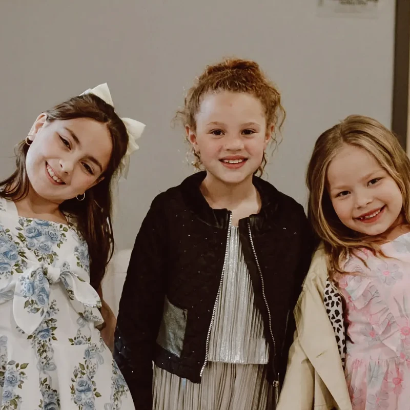 Three young girls in party dresses stand close together and smile at the camera. The girl on the left raises her arm cheerfully.