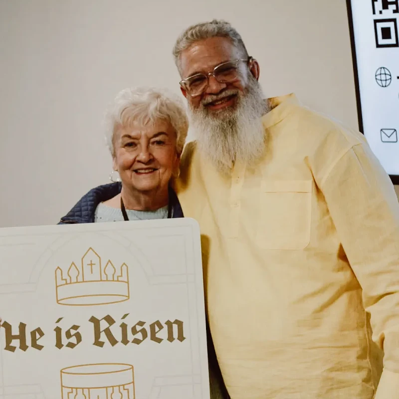 An older woman and a smiling man with a long white beard pose together. The woman is holding a white sign that says 'He is Risen' in gold letters.