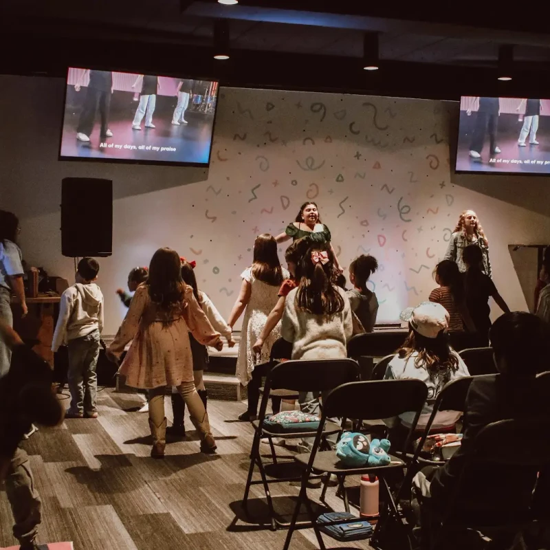 A group of children on a low stage singing and dancing, led by two women. Other children are seated watching, with screens showing song lyrics above.