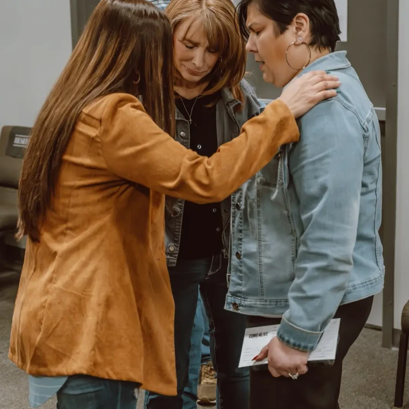 Three women stand close together in a prayer circle. One woman in a brown jacket places her hands on the other two women in a comforting gesture.