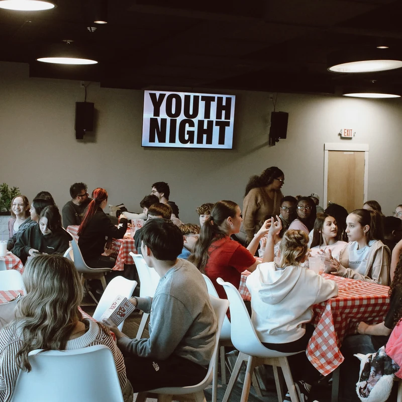A large group of teenagers sit at tables with red and white checkered tablecloths, talking and eating during a Youth Night event.