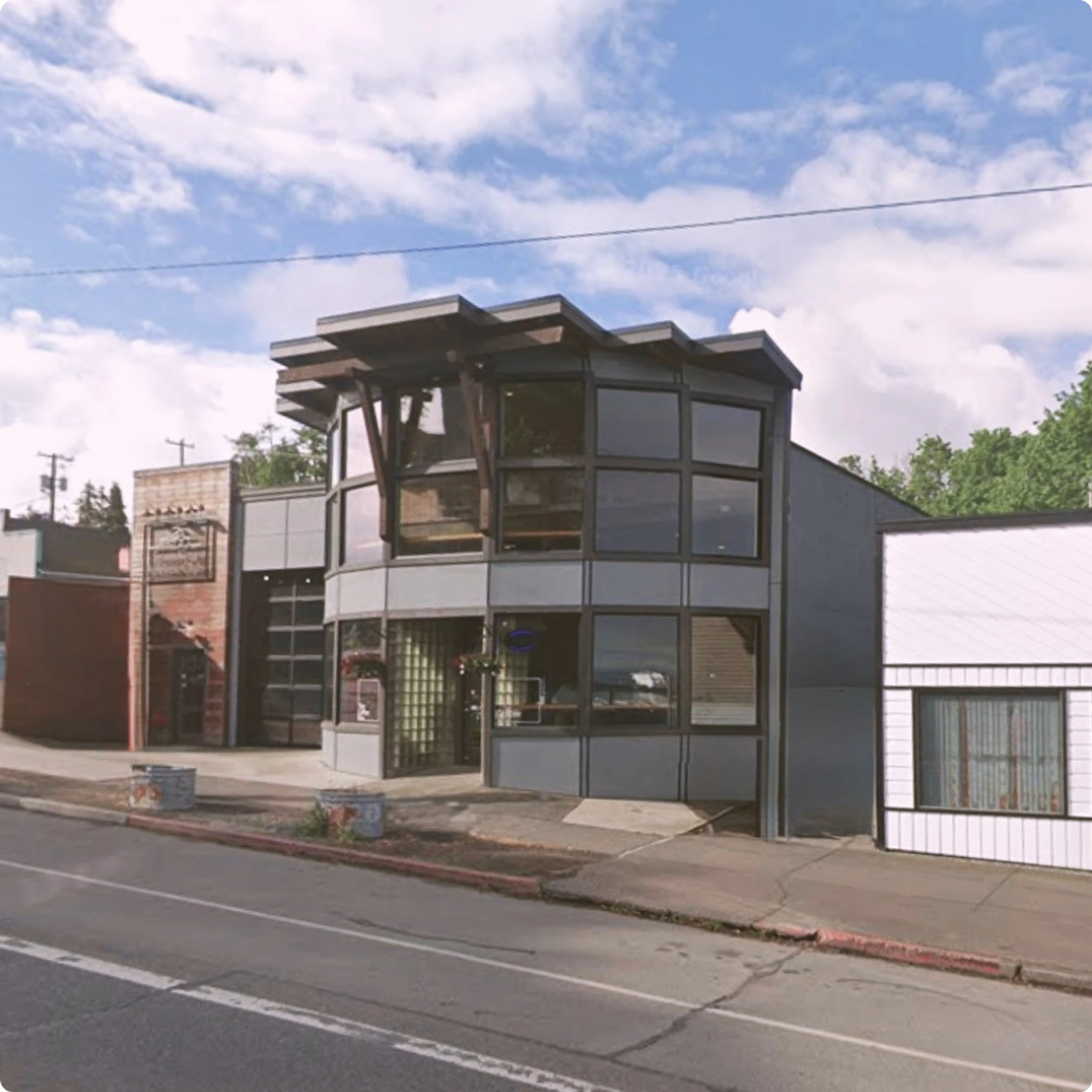 A modern two-story commercial building with a grey facade, large windows, and a distinctive wooden roof structure, situated on a street next to other shops.