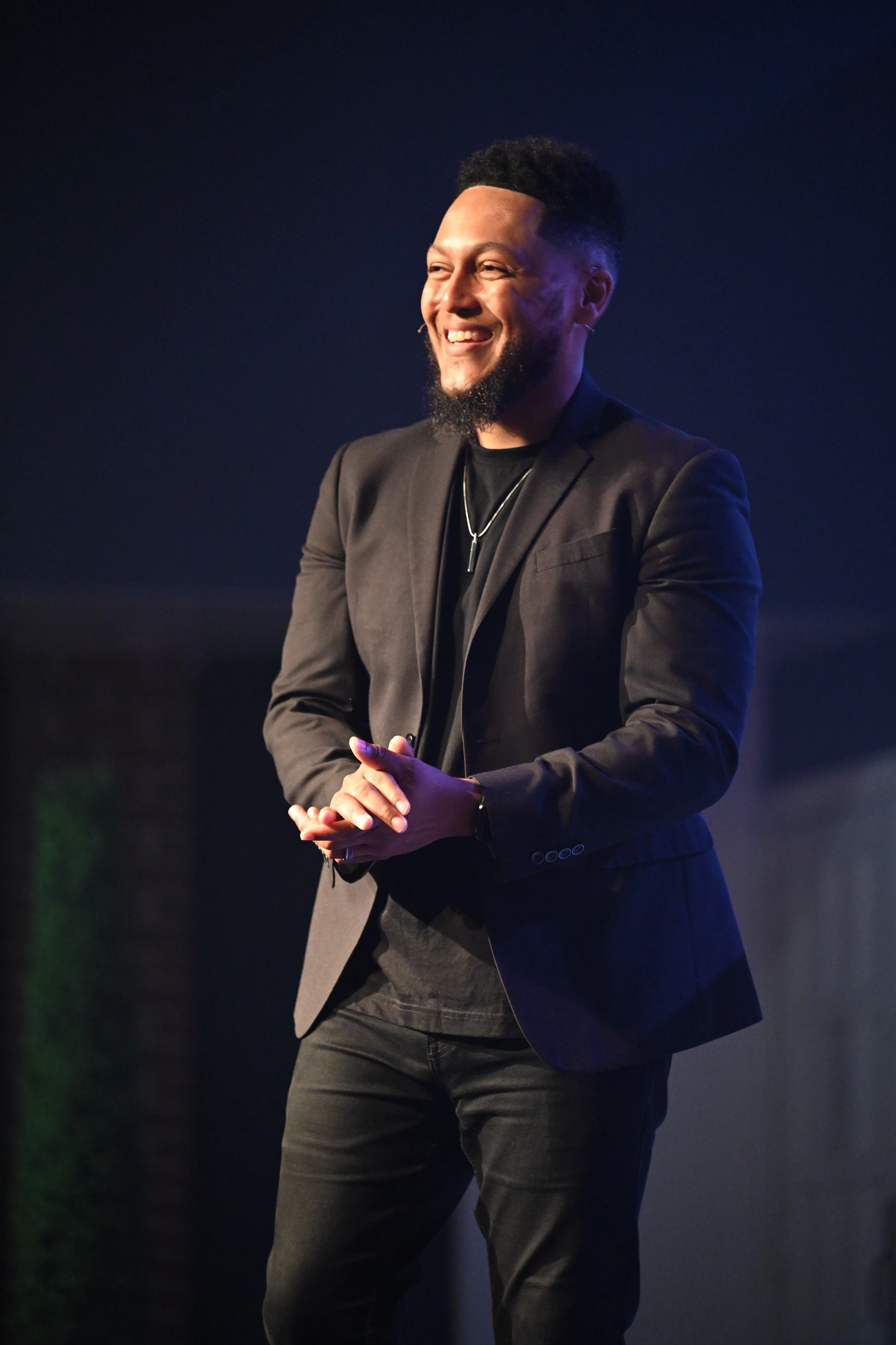 A smiling man with a beard, wearing a black blazer and t-shirt, stands on a dimly lit stage with his hands clasped, looking off to the side.