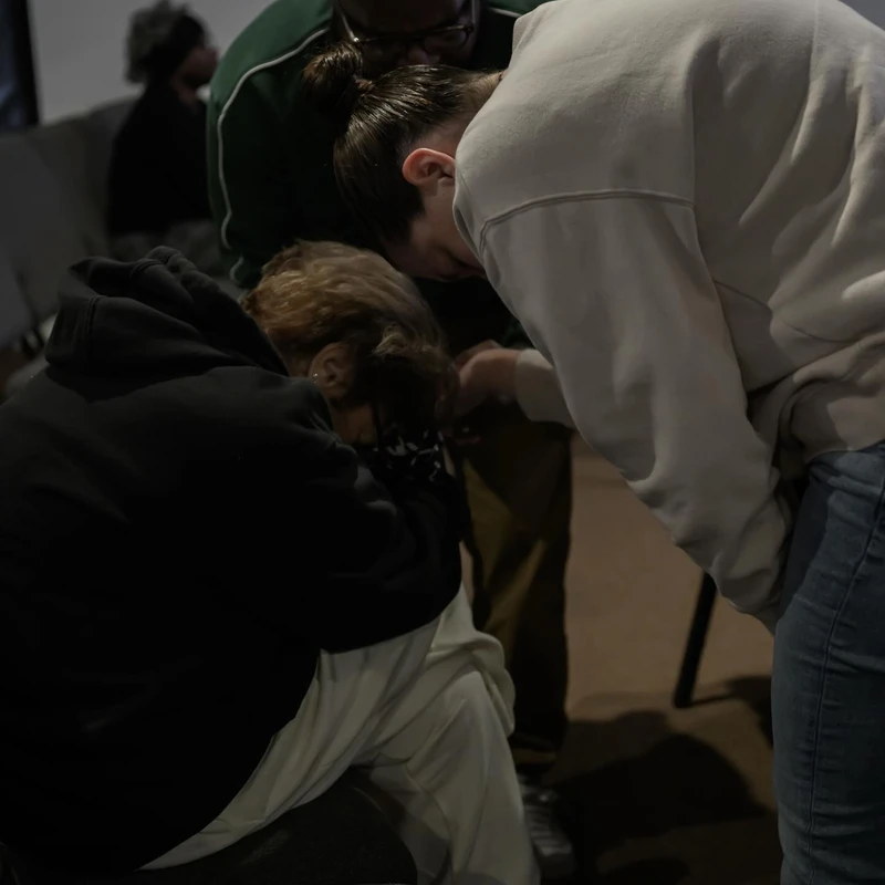 Three people gathered in a moment of prayer. One person is kneeling while a man and a woman lean over them with supportive hands in a dimly lit room.