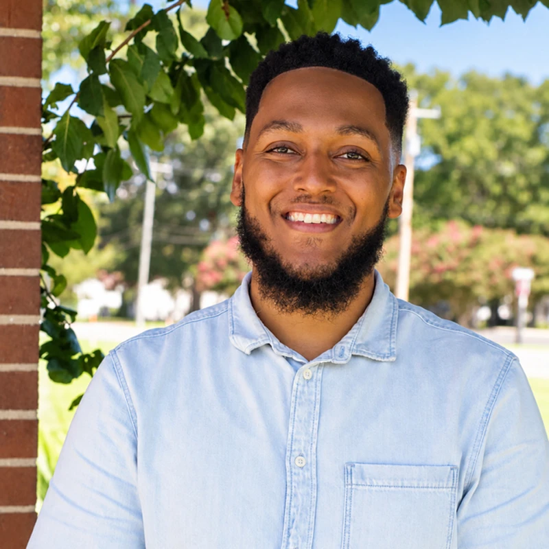 A smiling young Black man with a beard and short hair, wearing a light blue button-down shirt, stands outdoors on a sunny day with green trees behind him.