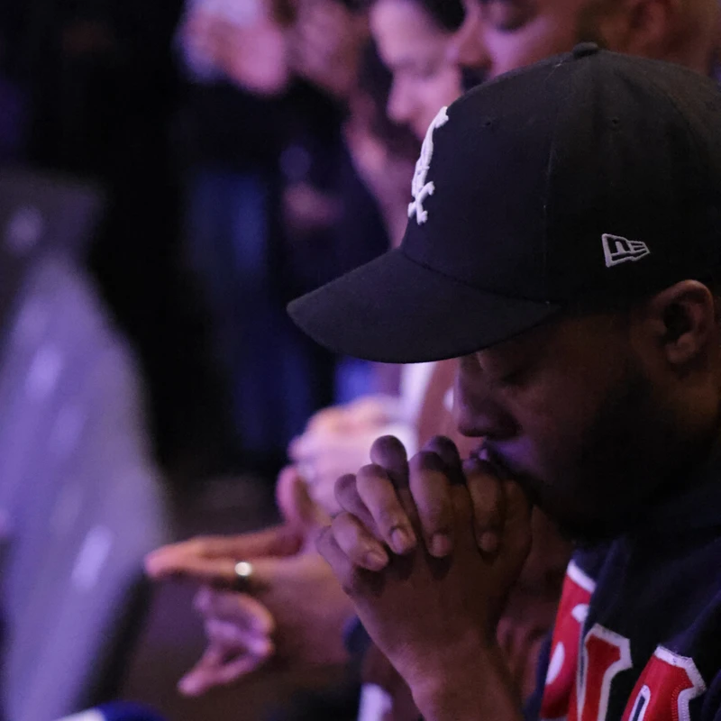 A close-up side view of a man in a baseball cap with his hands clasped in prayer. Behind him, a line of other people are also praying with eyes closed.