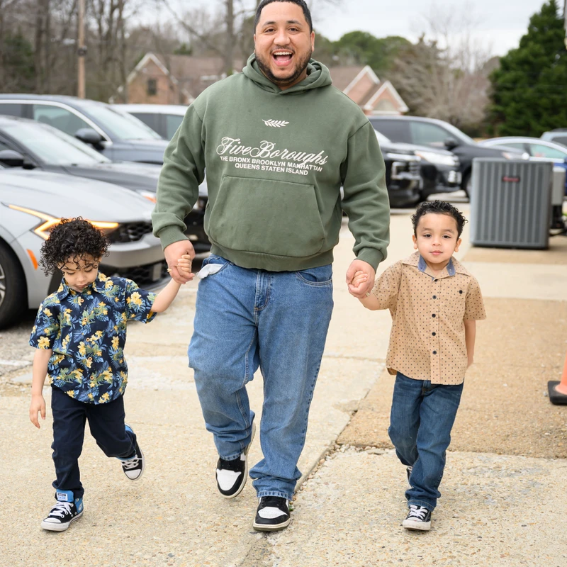 A smiling father walks toward the camera, holding hands with his two young sons on either side. They are in a parking lot on a cloudy day.