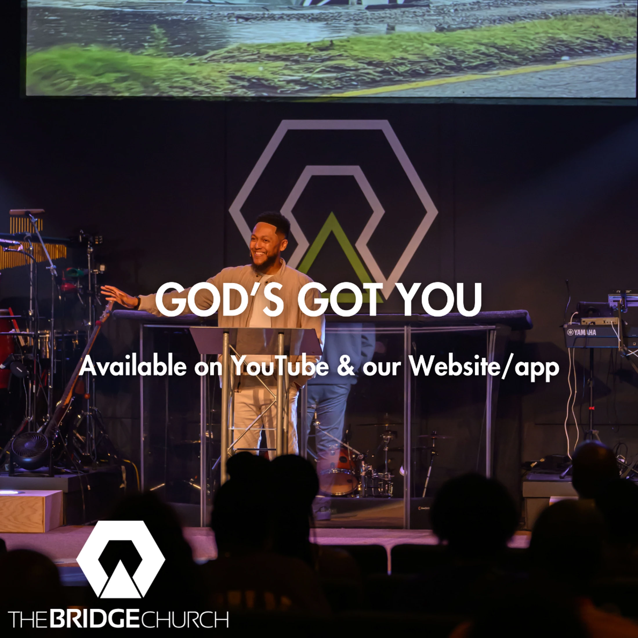 A smiling pastor stands on a stage behind a pulpit with text that reads "God's Got You, Available on YouTube & our Website/app" for The Bridge Church.