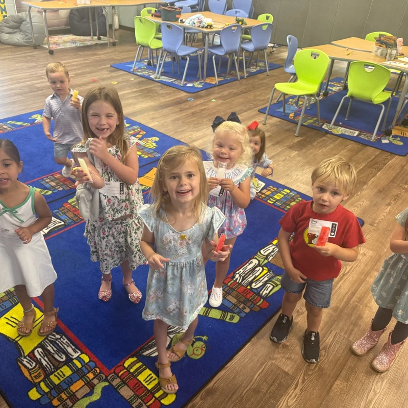 A group of young children in a classroom smile for the camera while holding colorful ice pops. Tables and chairs are in the background.