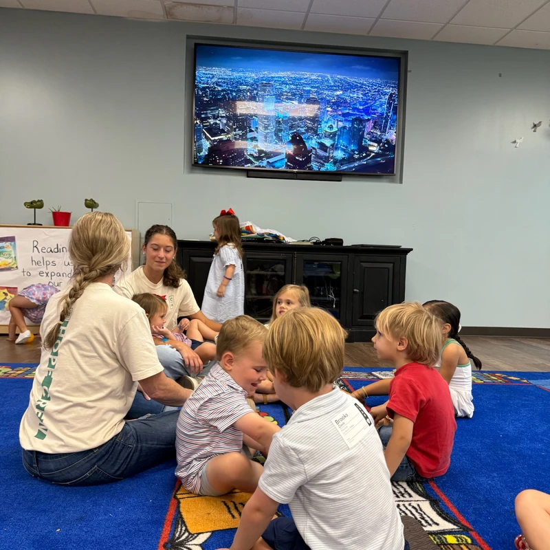 A group of young children and two women sit in a circle on a colorful blue carpet in a classroom, interacting during a group activity.
