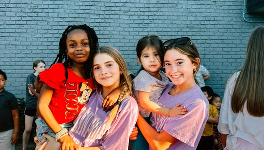 Four diverse young girls smile for the camera while posing in front of a blue brick wall. Two older girls hold two younger girls in their arms.