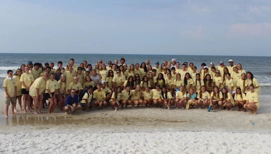 A large group of young people and adults in matching yellow shirts pose for a photo on a sandy beach, with the ocean and a clear sky in the background.